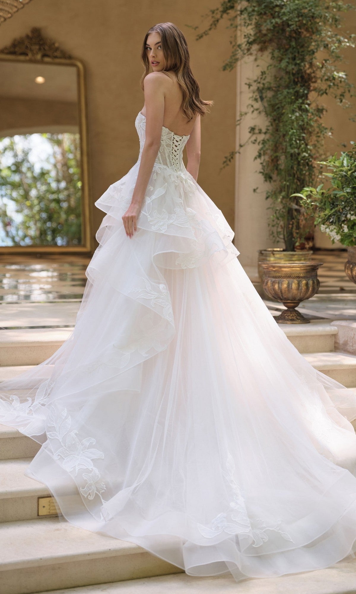 A woman in the Andrea and Leo WL059 Long White Wedding Dress with layered white tulle stands indoors on stairs, looking over her shoulder. Lush green plants and a large mirror are in the background.